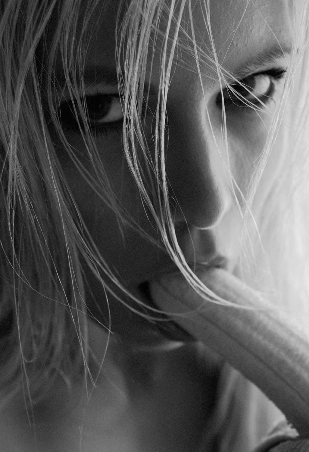Schwarz-Weiß-Fotografie eines Gesichts im Close-up. Die Person hat nasse, ins Gesicht fallende Haare. Eine Banane zwischen den Lippen.