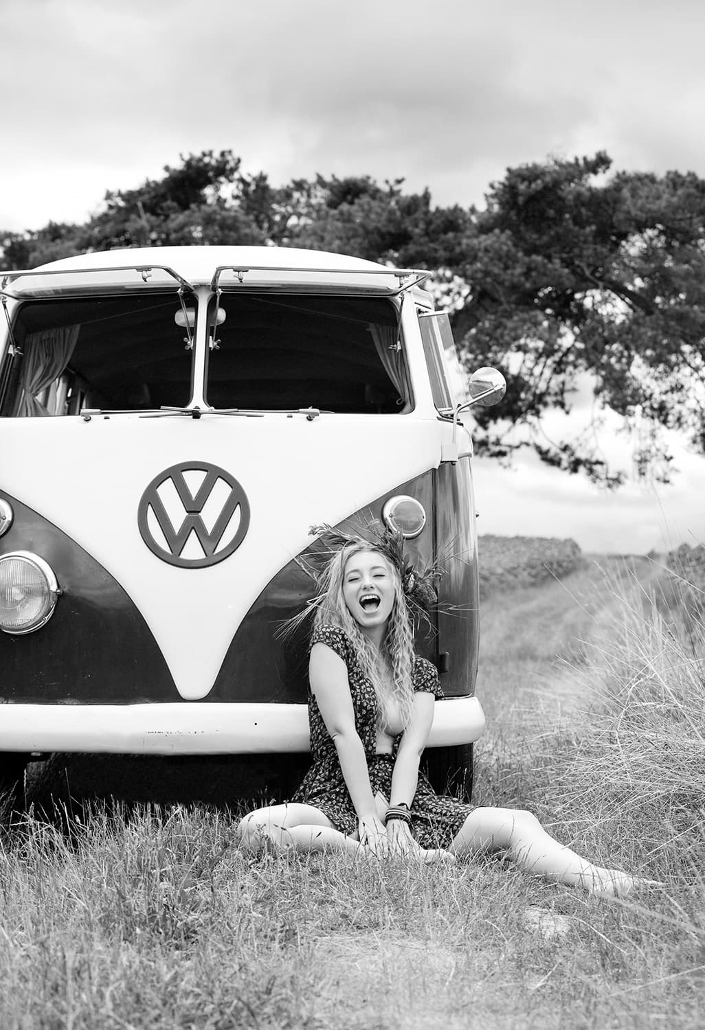 Schwarz-Weiß-Aufnahme einer Frau mit lockigem Haar, die vor einem Oldtimer-VW-Bus sitzt. Sie trägt ein leichtes Sommerkleid und lacht mit offenem Mund.