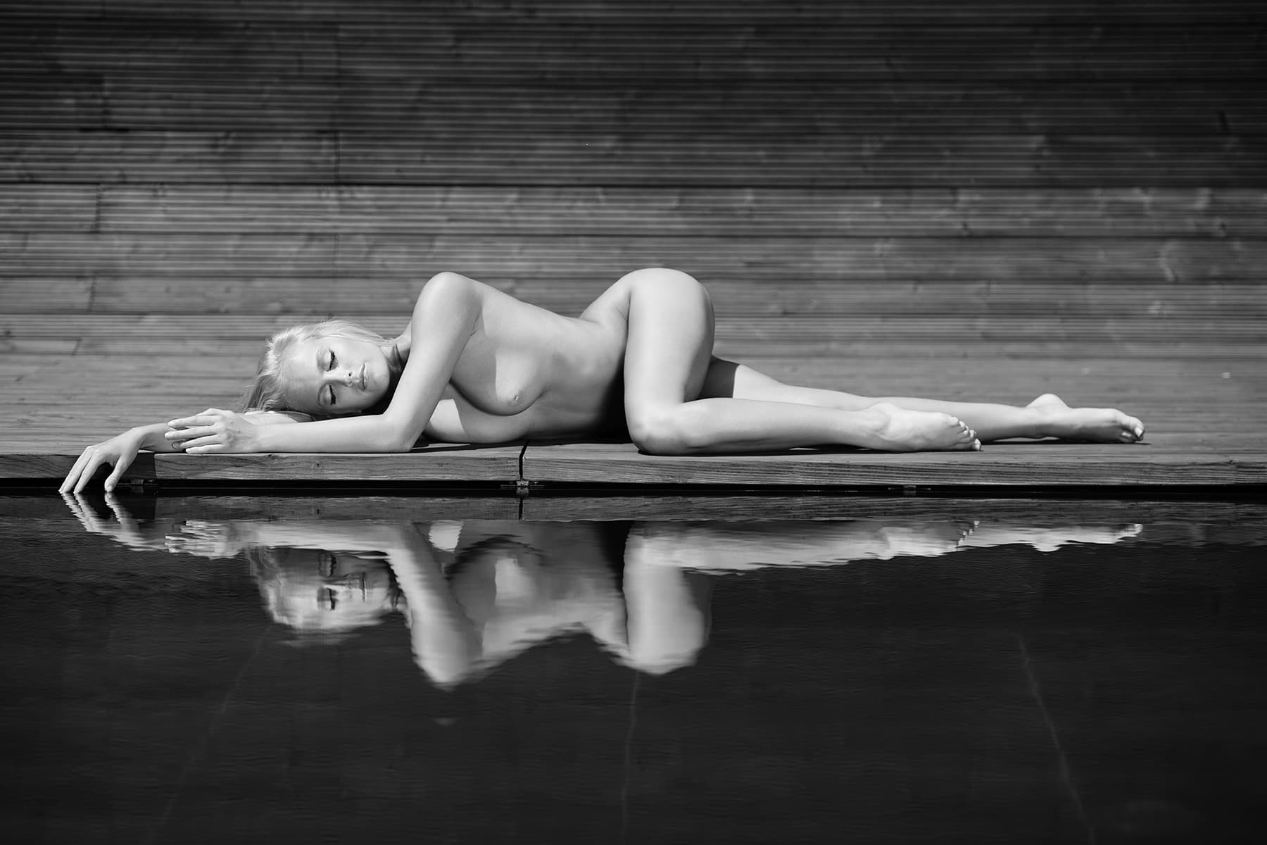 Limited edition fine art nude photography — woman lying on a wooden deck, reflected in still water. Black and white print by Martin Wieland.