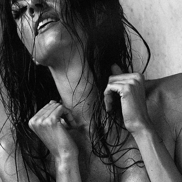 Erotische Fotografie einer nackten Frau, die sich leidenschaftlich hält, mit feuchtem Haar und intensiver Ausstrahlung in einem industriellen Raum.