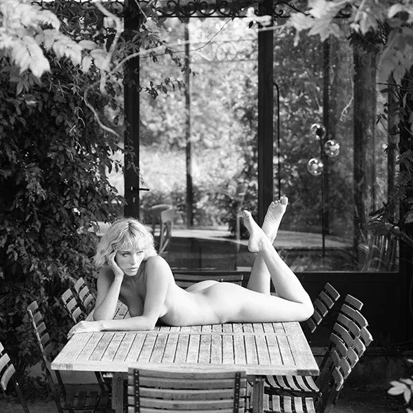 Nackte Frau liegt auf einem Holztisch, umgeben von leeren Stühlen, in einer atmosphärischen Schwarz-Weiß-Aufnahme - erotische Aktfotografie