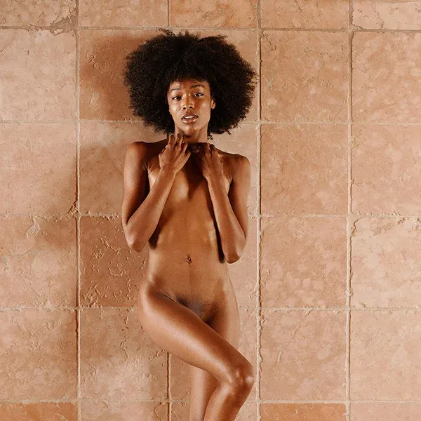 erotische Fotografie einer Frau vor einer Steinwand, ihre Pose ausdrucksvoll, mit einem natürlichen, klaren Blick. Die weiche Haut hebt sich von der rauen Mauer ab, die Schlichtheit der Komposition unterstreicht die puristische Schönheit des Körpers.