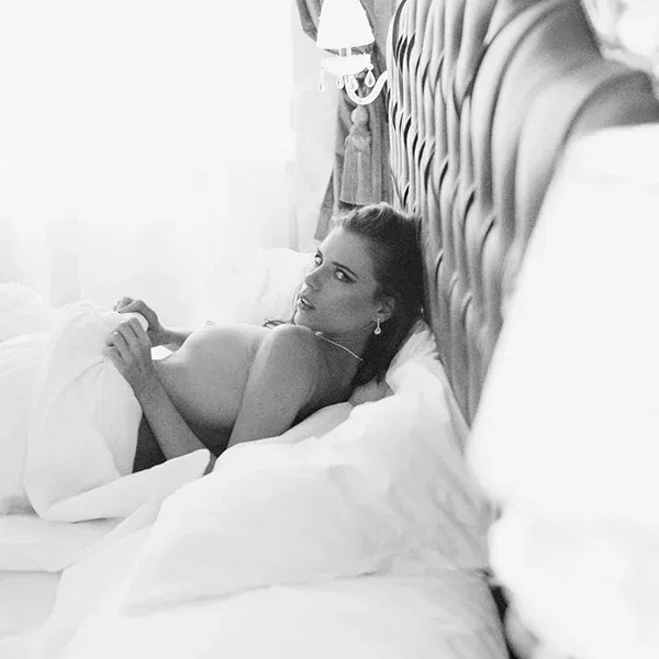 Erotische Fotografie: Eine verführerische Szene im Schlafzimmer, in der eine Frau mit sinnlichem Blick aus den weichen Kissen hervorblinzelt. Ihr nackter Oberkörper ist von der Bettdecke leicht bedeckt, und der Hintergrund mit den eleganten Lampen und Vorhängen schafft eine intime Atmosphäre.