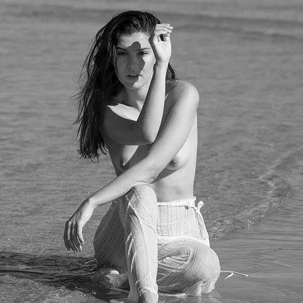 She-Came-for-the-Weather title Erotic photography on the beach - half-naked model kneeling in shallow water, direct view, arm for shade.